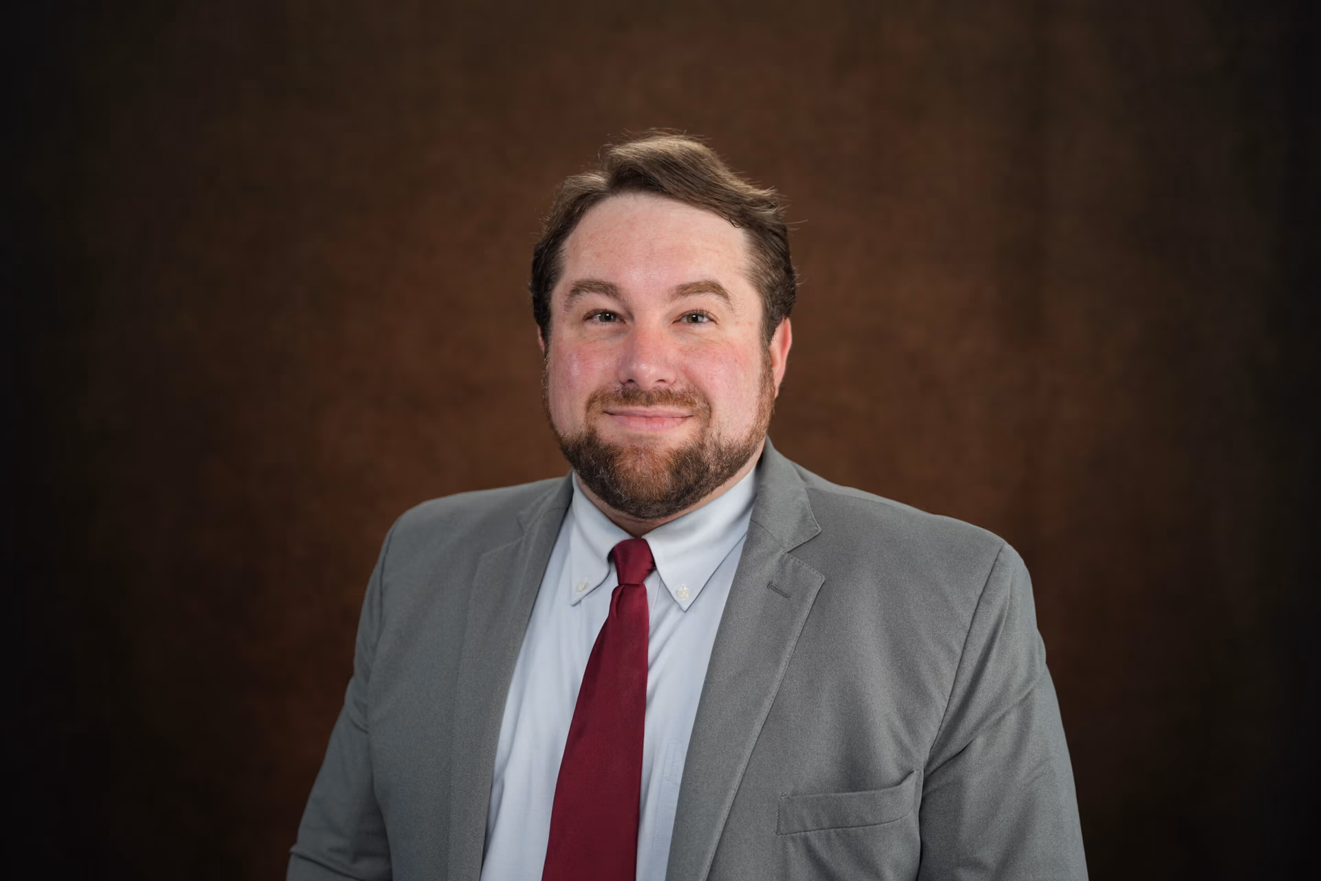 Professional portrait of a personal injury lawyer in a gray suit and red tie, smiling against a brown backdrop, representing Ethen Ostroff Law's commitment to client service and legal expertise.