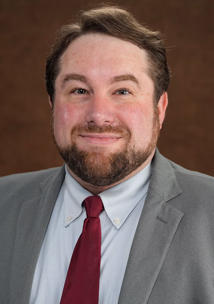 Brandon Zanan, personal injury lawyer at Ethen Ostroff Law, smiling in a professional gray suit with a red tie, against a warm brown background.